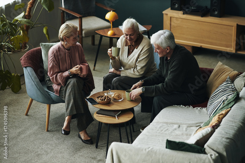 Three senior Caucasian women and man sitting together in living room drinking hot beverages and talking, snacks, cookies and biscuits on coffee table, relaxed social gathering among friends