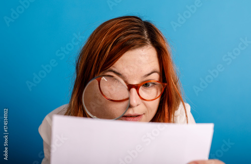 A woman with red hair, wearing glasses, and using a magnifying glass to examine a document, set against a vibrant blue backdrop. This image capt