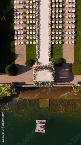 drone view of luxury outdoor wedding ceremony on Lake Como with floral arch, rows of white chairs on lush green lawn, and drone flying back to reveal panoramic lake and mountain views