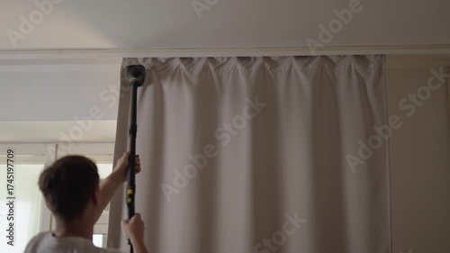 Rear view of housekeeper using handheld steam iron to smooth out wrinkles and creases from curtains hanging in a home, representing the concept of efficient textile care and domestic productivity.