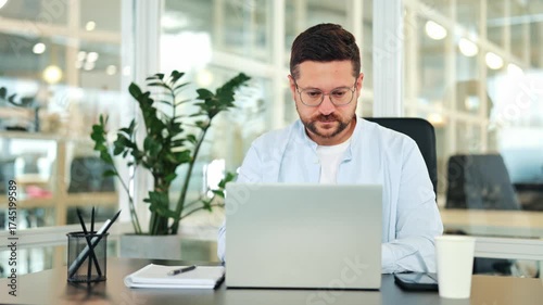 Focused businessman in a light blue shirt put on glasses works diligently on a laptop at a modern office desk. Professional specialist appears concentrated, managing tasks and operations efficiently.