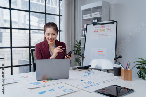 Smiling businesswoman connecting online using a laptop and smartphone, holding a pen during a virtual meeting, surrounded by business charts and a whiteboard in a modern office setup