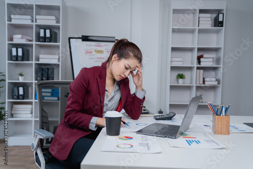 Asian businesswoman suffering from severe headache and stress, taking a break from work at her office desk, feeling exhausted and overwhelmed by the demanding workload and project deadlines