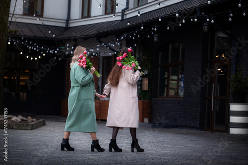 Two women walking together in stylish coats, holding vibrant bouquets of flowers, enjoying a cheerful moment outdoors, showcasing friendship and joy in a lively urban setting