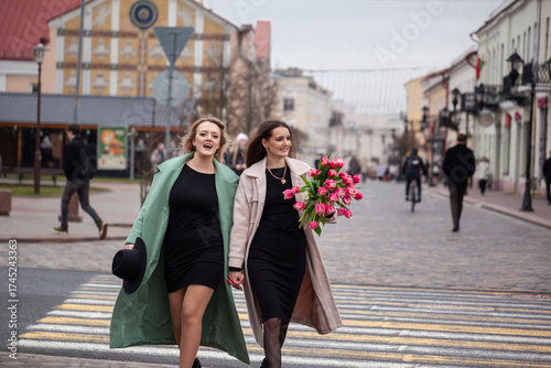 Two young Caucasian women walk together on a city street. One wears a green coat and the other a pink coat, both smiling and enjoying their time. One holds a bouquet of flowers