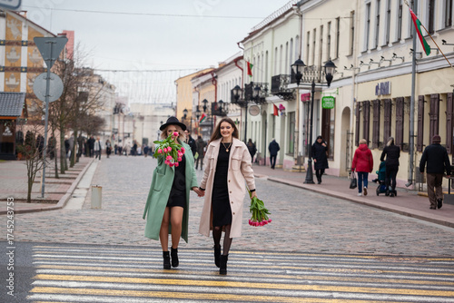 Two women walking hand in hand on a cobblestone street, dressed in stylish coats, one holding a bouquet of flowers, showcasing friendship and urban lifestyle