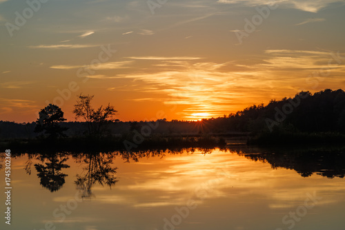 Golden sunset glows above calm Pine Barrens lake, with trees reflecting on still water beneath a warm orange sky at the end of the day.