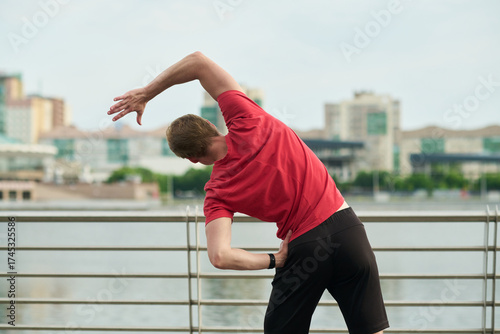 Young man stretching arms and torso outdoors near waterfront standing with back to camera performing side bend exercise with city buildings in background