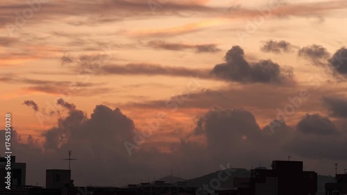 A vibrant sunset with orange and purple hues over a city skyline. Silhouettes of buildings and clouds create a dramatic scene.
