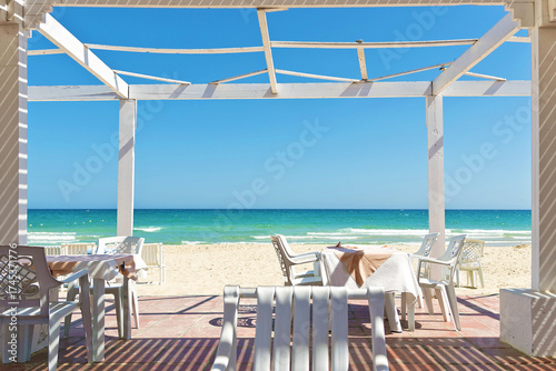 White chairs and tables in wooden pergola on a sandy beach, clear blue sky and calm sea in Tunisia. Gazebo or patio is place for relax on sea shore in restaurant 