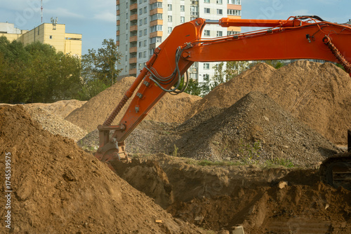 Excavator digging soil on construction site with urban buildings in the background. Industrial excavator performing earthmoving operations at construction site for residential infrastructure.