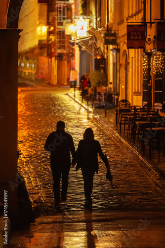Fototapeta Couple walking on a wet, cobblestone street with bars and restaurants at night i