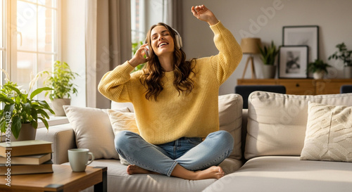 Joyful young woman blissfully dancing to music with headphones on a cozy couch, bathed in warm sunlight.