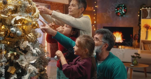 Cheerful grandparents and two little granddaughters decorating Christmas tree together. Joyful multigenerational family bonding during festive holiday season at home.