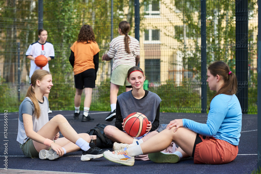 Fototapeta premium Several happy restful teenage girls sitting on sports court talking and relaxing after streetball game against their friends playing basketball