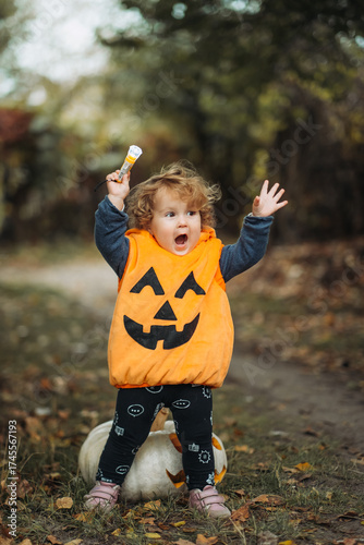Cheerful child in pumpkin costume standing in autumn forest on Halloween.