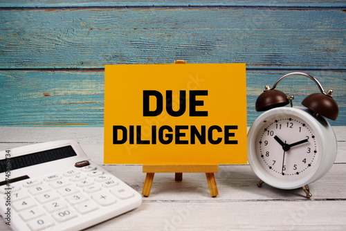 Due Diligence text written on paper card with calculator and alarm clock on wooden background