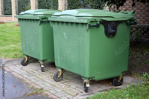Two green wheelie bins stand ready for trash collection on a paved area near a building, representing municipal waste management and local environmental cleanliness outdoors.