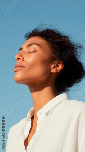 Woman enjoying sunlight, eyes closed, with a clear blue sky background.