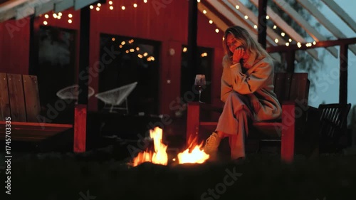 A woman sits by a fire in autumn against the backdrop of a house with garlands