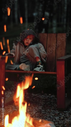 A boy sits in a garden chair near a campfire in nature, vertical video
