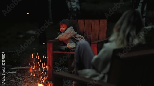 A boy sits in a chair by the fire and talks with his mother