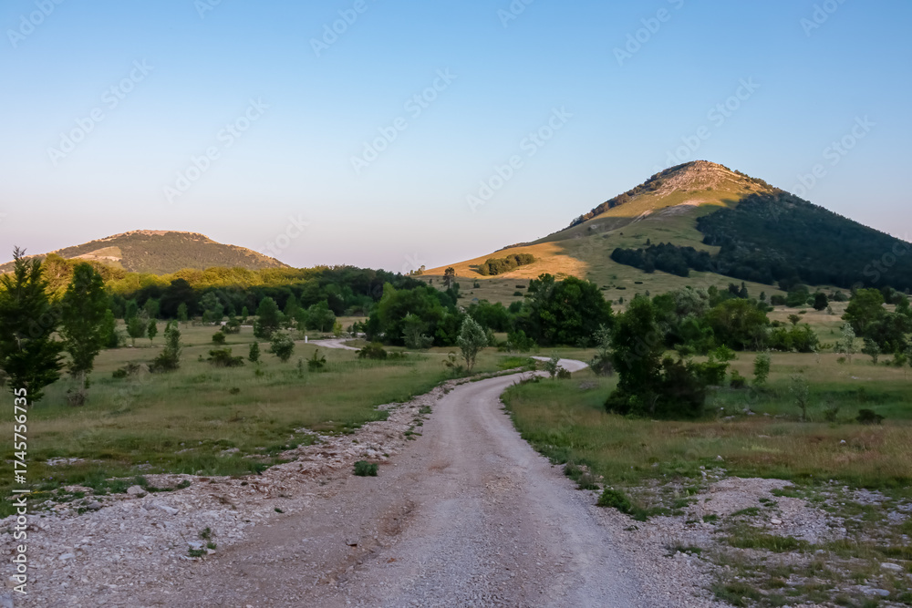 Fototapeta premium A winding gravel road meanders through a serene valley in Croatia's Velebit mountains, leading towards the sunlit peak of Kiza under a clear blue sky during a peaceful summer evening.