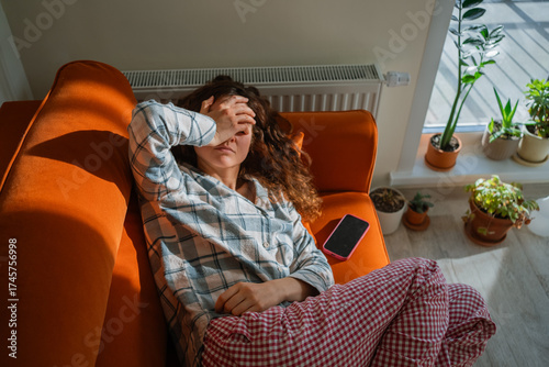 Woman feeling unwell and exhausted while lying on an orange sofa, covering her eyes with a hand, suffering from toxicosis, nausea, or a headache, indicating discomfort and illness