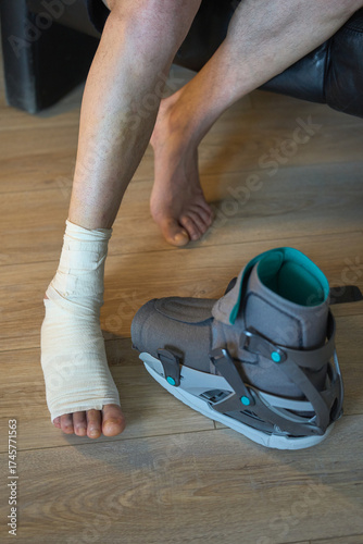 Bandaged foot and orthopedic boot on wooden floor. Injured foot with bandage next to orthopedic boot. Recovery in progress.