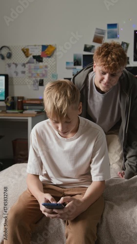Vertical shot of young teenage boy sitting on bed and typing on smartphone with older brother coming up behind him and stealing phone teasing
