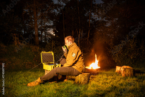 Amateur radio operator outdoors by a campfire, operating portable HF radio from a Go-Box in the wilderness, enjoying nature, emergency communication and Parks on the Air activity