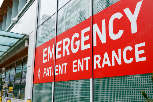 Bold red emergency patient entrance sign on modern glass hospital facade, with walking icon and drop-off zone amid urban setting