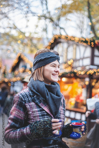 Beautiful woman holding two cup of mulled wine, while standing on traditional Christmas market in Germany. Portrait of cheerful model in winter outfit social smiling with cup of tea or coffee.