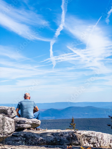 Man sitting on rocky outcrop overlooking wide mountain landscape and dramatic sky with white contrails at Bear Rocks Preserve, Dolly Sods, West Virginia