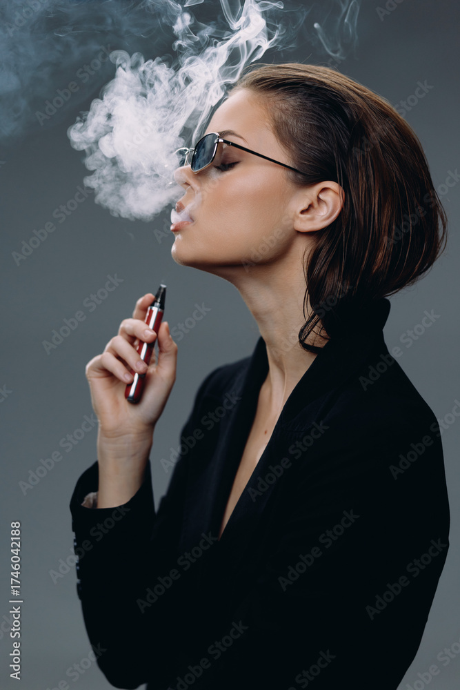 Fototapeta premium Young woman vaping with smoke in a dark studio, wearing sunglasses and black jacket, stylish and modern.