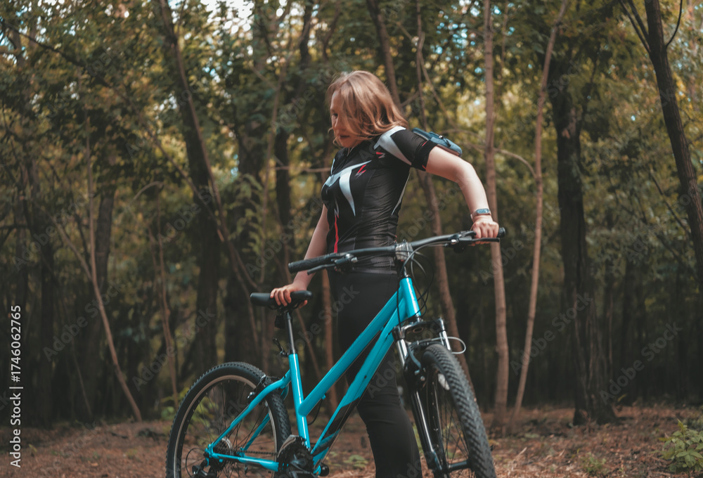 Obraz premium Smiling woman in sportswear pauses with bicycle in a serene forest, embracing a healthy lifestyle and enjoying nature on a sunny day