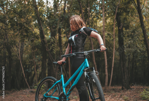 Smiling woman in sportswear pauses with bicycle in a serene forest, embracing a healthy lifestyle and enjoying nature on a sunny day