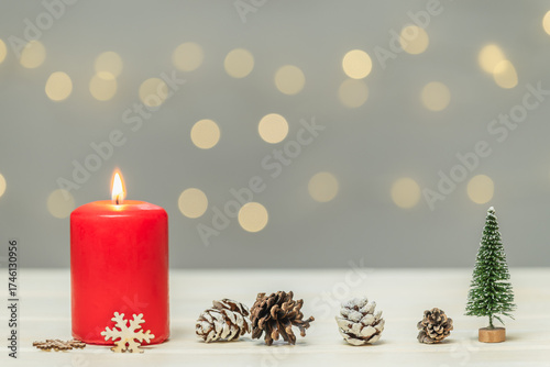 A red burning candle, pine cones, and a small artificial fir tree against a blurred background with bokeh. Christmas background, greeting card.