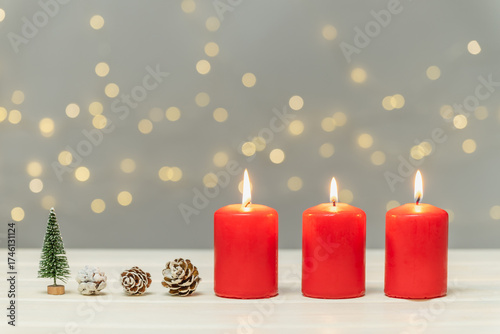 Three red burning candles, pine cones, and a small artificial fir tree against a blurred background with bokeh. Christmas background, greeting card.