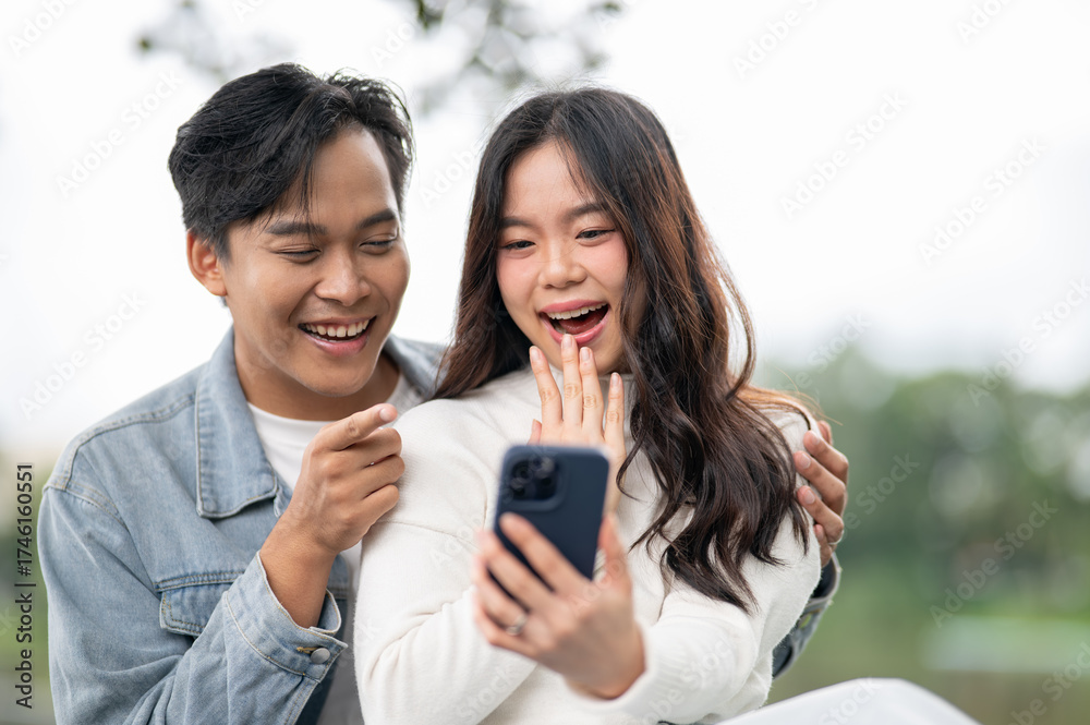 Fototapeta premium Young asian couple looking at phone a man hugging woman's shoulder while sitting outdoors in park.