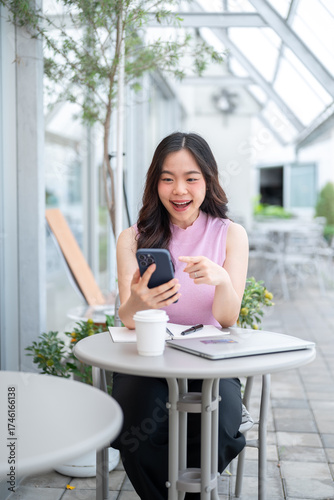 Asian woman student holding and laughing at phone while sitting with laptop and coffee at cafe table Free Download Asian woman student holding and laughing at phone while sitting with laptop and coffee at cafe table - GFXLoad