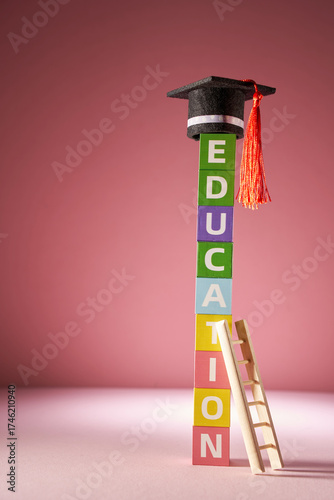 small mortarboard on top of colorful toy blocks with single word education and ladder against pink background.