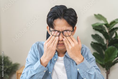 Eye problem concept, fatigue asian young man tired eyes, irritated rubbing eye dry relief vision pain after work on laptop computer, eye strain, while wearing glasses, suffer migraine, bad blurry.