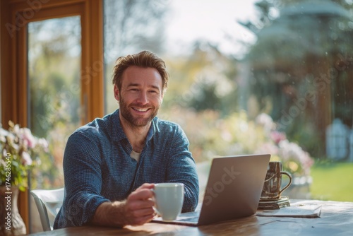 A happy man sitting at a table, using a laptop in a bright, modern home office. the man he is smiling while working online or looking for inspiration.