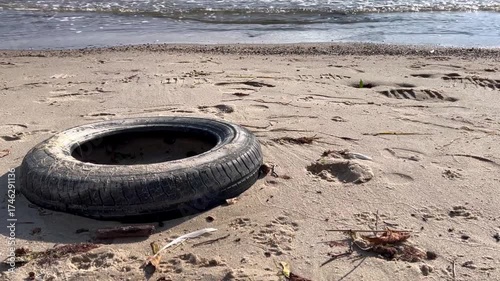 An old black rubber tire lying on a sandy beach with sea waves in the background. Marine pollution and environmental issues caused by abandoned waste on coastal areas