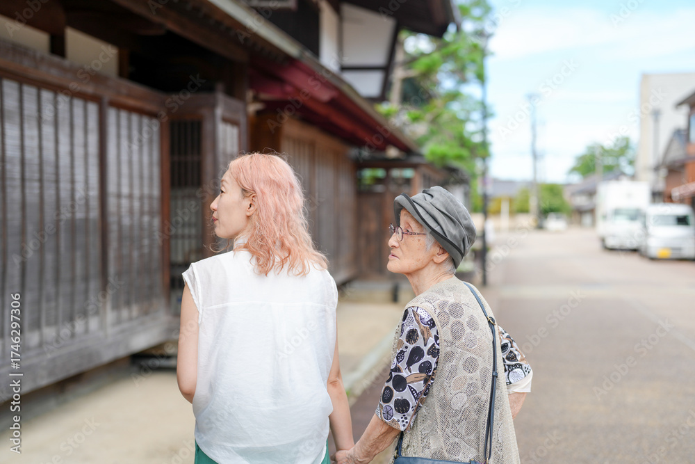 Fototapeta premium Two women walking down a street, one with pink hair. The woman with the pink hair is holding the hand of the older woman
