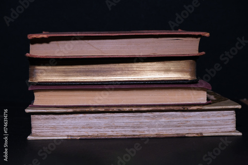 Side view of a pile of antique books arranged carefully on a dark surface, showing textured bindings, faded titles and vintage paper tones, ideal for education or historical themes.
