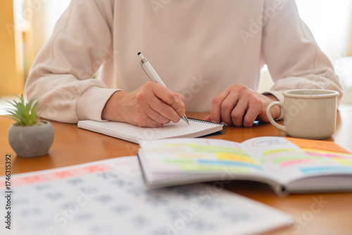 Woman's hands writing notes in a physical planner or journal, organizing daily activities and appointments for improved productivity and a balanced lifestyle at a home office desk