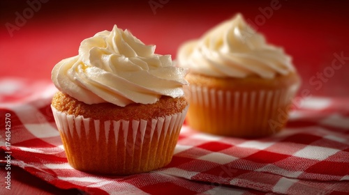 Two delicious vanilla cupcakes topped with creamy white frosting, resting on a red and white checkered cloth.