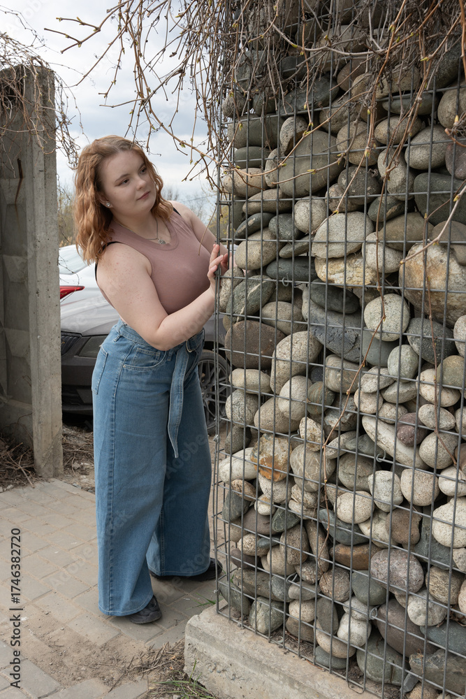 Fototapeta premium Young lady standing beside a wire mesh gabion with rocks. Modern landscape design element.